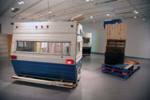 Two blue and white trailers sit in a gallery space. A wooden skid with tires and miscellaneous construction material on top sits to the right of the trailer.