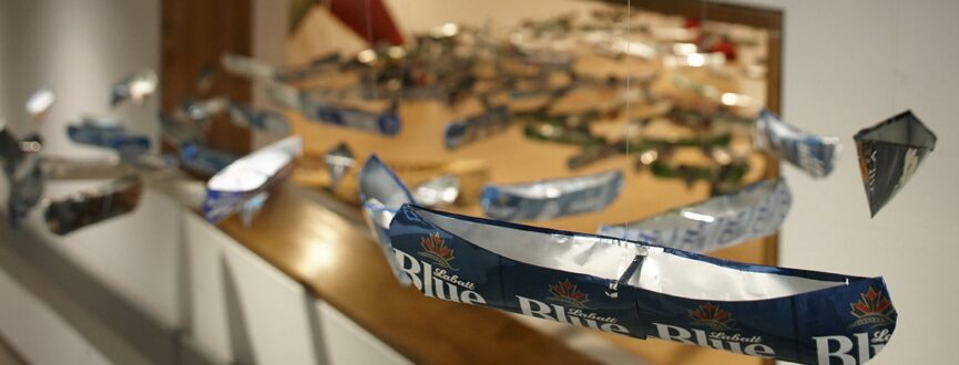 close up view of a small canoe made from a Labatt Blue aluminum beer can and suspended from the ceiling. Hundreds of other canoes made from other brands of beer are visible out of focus in the background