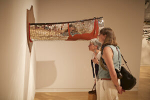 two people view an installation based artwork of a mirror mosaic canoe cut in half and mounts on a plaque on a gallery wall