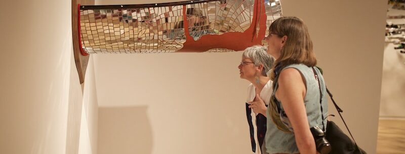 two people view an installation based artwork of a mirror mosaic canoe cut in half and mounts on a plaque on a gallery wall