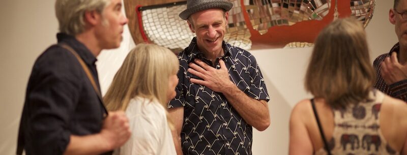 Brad Copping stands in front of one of his sculptural installation pieces with a hand on his chest, smiling at a gathering of people around him