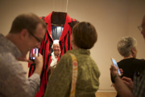 A person enveloped in a very large art installation that is made to look like a jacket in red and black stripes. In the foreground are people that are slightly out of focus who are taking a photo of the person in the coat