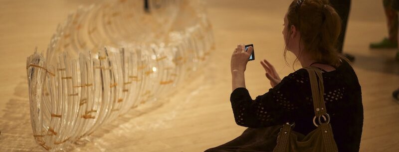a person sitting on the ground and holding up their phone on the right to take a photo of a glass canoe on the left