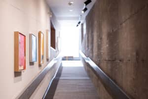 paintings hanging in an art gallery hallway on the left and a concrete wall on the right. The hallway extends up toward a window at the end