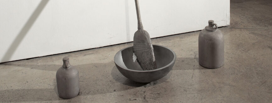 Cast iron sculpture in grey of two jugs (in cast iron) flanking either side of a straw broom et in a bowl, also case out of iron. The sculpture sits on a concrete floor in front of a white wall.
