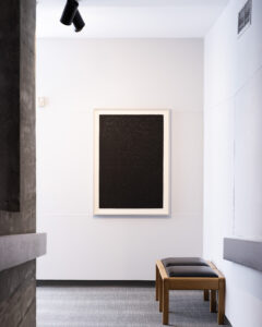 Installation view of the lower ramp of the Art Gallery of Peterborough, featuring a Tim Whiten print hanging on the wall. Two wooden seats sit along the right wall. To the left, the concrete divider and railing are visible