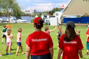 two art gallery volunteers wearing red shirt supervise campers playing outdoors in del crary park facing the river