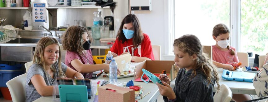 Students sit around a studio table creating stop motion animation figures out of clay while a gallery volunteer in a red t-shirt assists and supervises