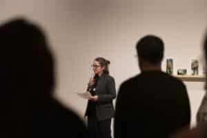 Fynn Leitch holds a microphone up to her face and holds a sheet of paper as she delivers opening remarks at an exhibition. Two figures appear silhouetted in the foreground and Leitch appears in the gap between them