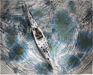 A white and black kayak adorned with antlers and stone is mounted on top of a canvas textile that is printed with the striations of several a tree ring.