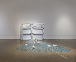 Two white shelves open up to a cascade of beaded threads, transitioning from silver to blue. On top of the beaded threads sits a silver beaver holding a book. The beaver faces out toward a gathering of four ducks that face toward it.