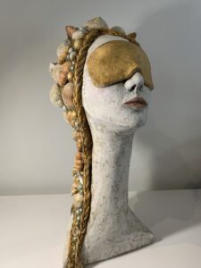 Sculpture of a head and neck with long gold braided hair with shells intertwined from the crown of the head down to the neck. A golden eye mask covers the eyes, the figure looks relaxed.