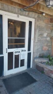 The exterior of the artist's studio; a while screen door with two signs that read "Open" and "Welcome"