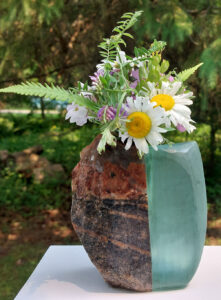 The artist’s work displayed in the outdoors on a white plinth. A vase that is half glass half stone holds fresh cut wildflowers.