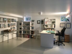 The interior of the artist’s studio; paintings are hung on the walls and displayed on shelving units on stands. There is a work desk and chair.