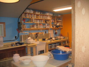 The interior of the artist’s studio; counters with various materials and tools, a small collection of blue mugs, and white bowls.