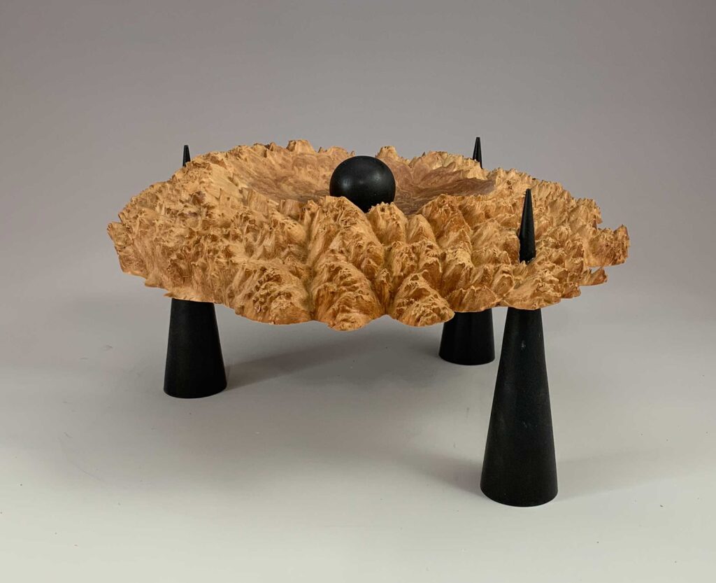 A highly textured piece of wood, with a black ball sitting in the centre, is supported off the ground by three black cones that penetrate the wood on three sides.