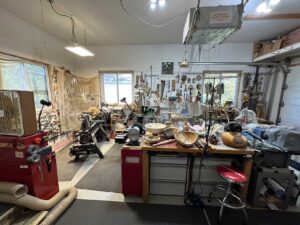 The interior of the artist’s studio; woodworking shop with tools hanging on the walls, a lathe, and other large equipment.