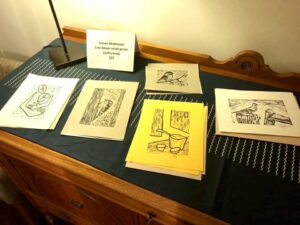 The interior of the artist’s studio; a table arranged with a selection of artist works on paper with a sign that says “James Matheson Lino block relief prints (unframed) $25”