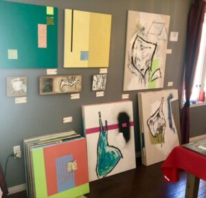 The interior of the artist’s studio; a wall arranged of the artist’s works on canvas, each with a title and price card.
