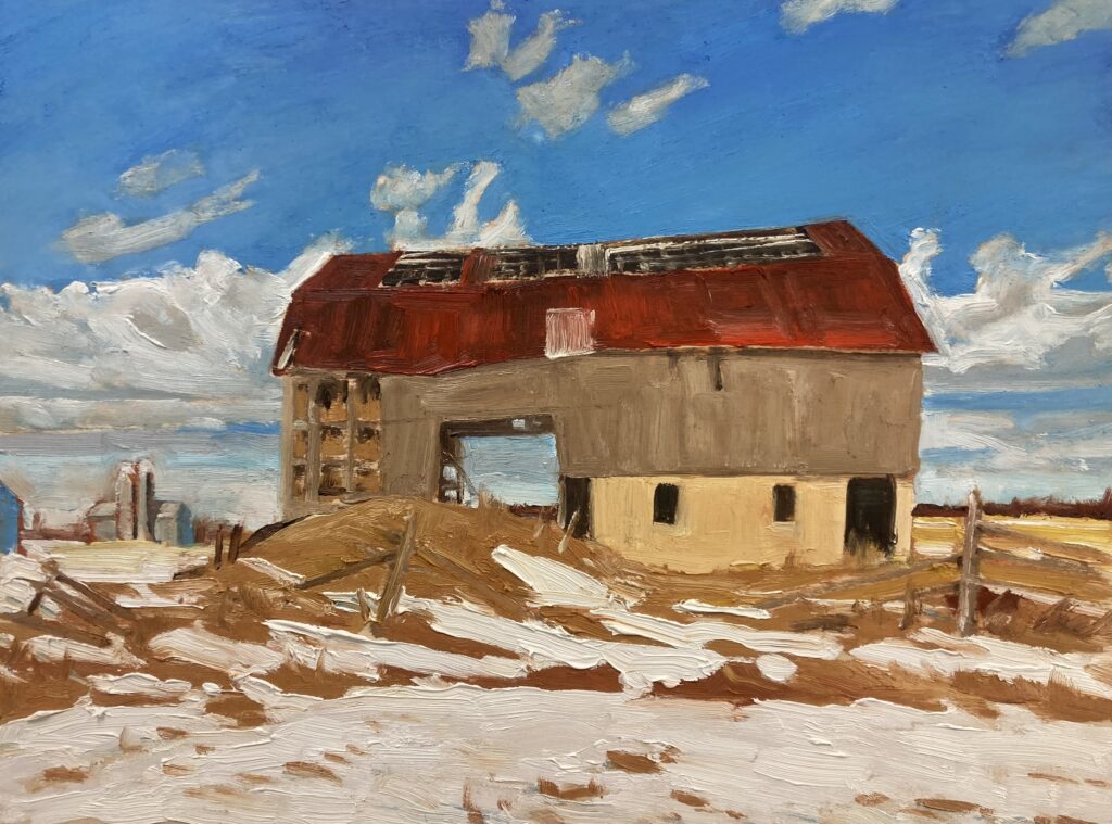 A weathered wood barn with a red roof is centred in a field partially covered with snow, it sits under a blue sky with clouds. A farm building can be seen in the distance on the horizon line.