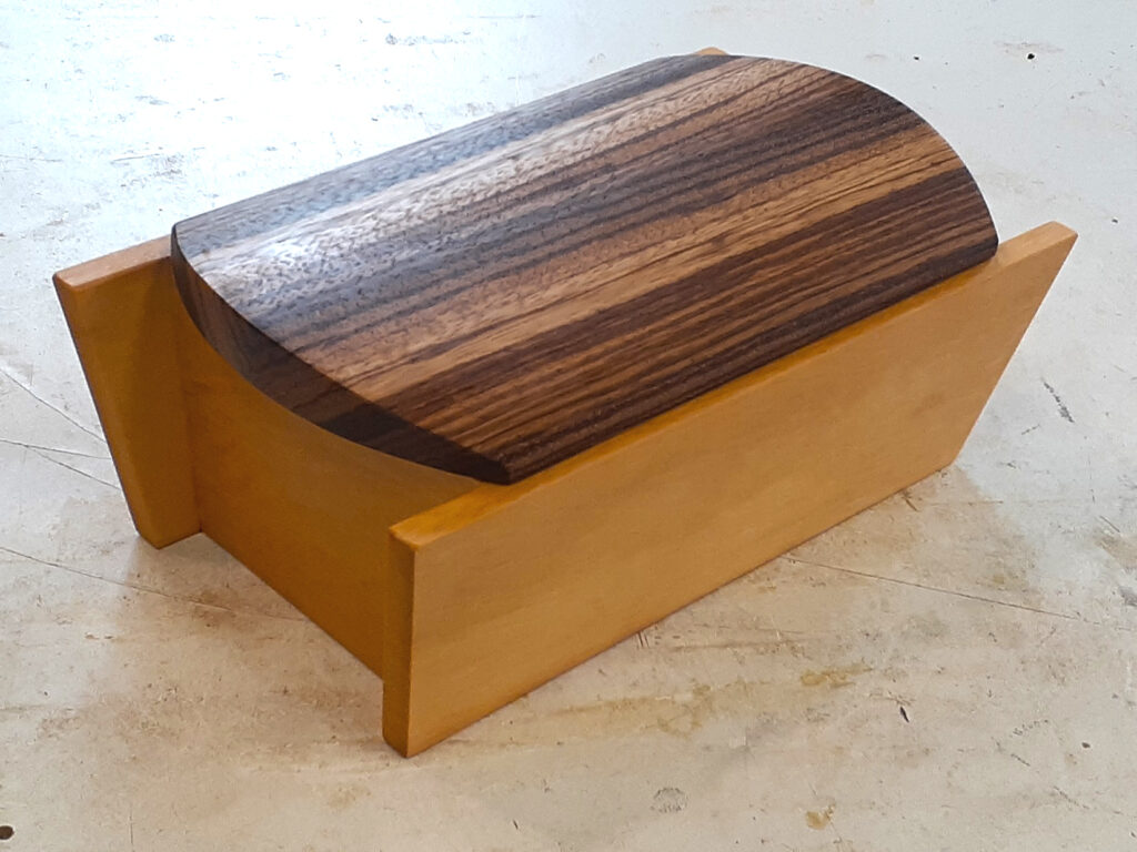 A closed wooden box; the top is made of a dark wood with a distinctive wood grain. The base is made from a lighter wood that forms a square box with the horizonal sides extending beyond the vertical sides.