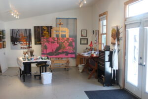 The interior of the artist’s studio; a large canvas set on an easel with tables near by holding paint and brushes. There are works displayed on the walls of the studio.