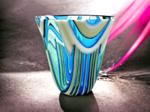 A blue and white glass blown vessel with a geometric line patterning. The vessel is sitting on a black surface