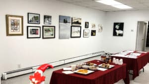 Interior of the Art school of Peterborough; paintings and drawings are displayed on a white wall. Two tables, with red table clothes, are in the middle of the room with food and drinks on them.