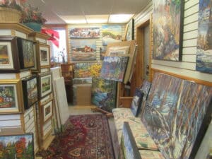 The interior of the artist’s studio; many landscape paintings by the artist are arranged on the walls, some are framed and some are unframed. A tall easel shows additional works.