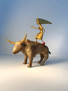 A gold human figure with an irregular shaped head, wearing red shoes stands on the back of an gold bull with horns.