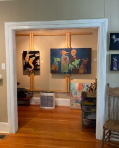 The interior of the artist’s studio; two of the artist’s works on canvas are on display on a wall; they are framed by the white door trim from the adjoining room.