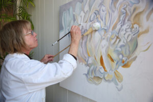 The artist working with two painting tools on a flower piece with mute tones. The work is unfinished on a large canvas.