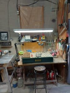 The interior of the artist’s studio; a work bench with tools arranged on the wall, a overhead light, and a stool.