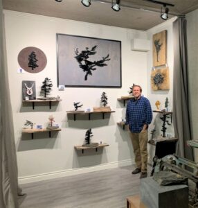 The interior of the artist’s studio; the artist is standing beside a wall display of their works on floating shelves as well as large hanging works.