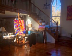 The interior of the artist’s studio; a large easel with a canvas work on it and tables beside it. Paint tubes, paint brushes, and water containers are on one table; a lamp, papers, and small paintings are on the other table. There is a large window and wooden staircase leading up to a second-floor loft