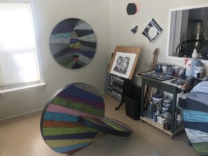 The interior of the artist’s studio; a white room with a colourful sculpture in the centre. There is a cart of the art supplies and other works beside the sculpture