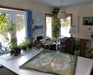 The interior of the artist’s studio; a large working table with a textile piece on it. There are tools and materials arranged around the room as well as a sleepy cat and some house plants.