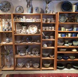 The interior of the artists’ studio; a wooden shelf display of the artists’ ceramic work: mugs, bowls, plates, vases.