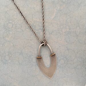 An abstract shaped pendant; a pointed bottom with a half circle fixed to the chain with three hoops. The work is displayed on a light blue patterned background.