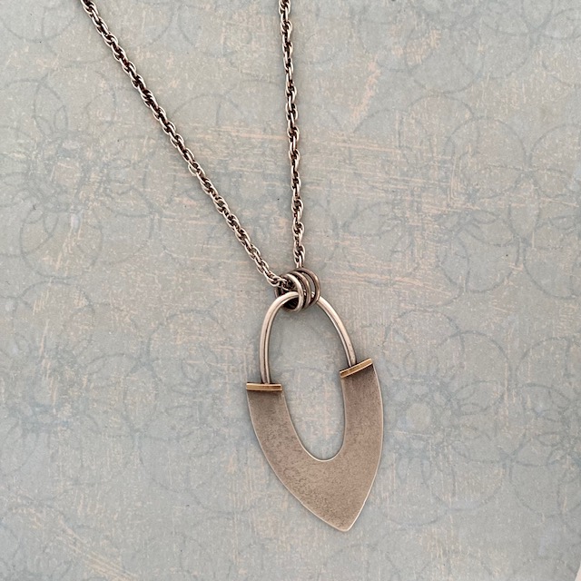 An abstract shaped pendant; a pointed bottom with a half circle fixed to the chain with three hoops. The work is displayed on a light blue patterned background.